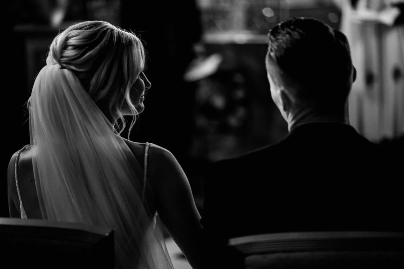 A bride glances at her groom during a quiet moment in their church wedding ceremony - powerful black and white wedding photography by Oben Studio®